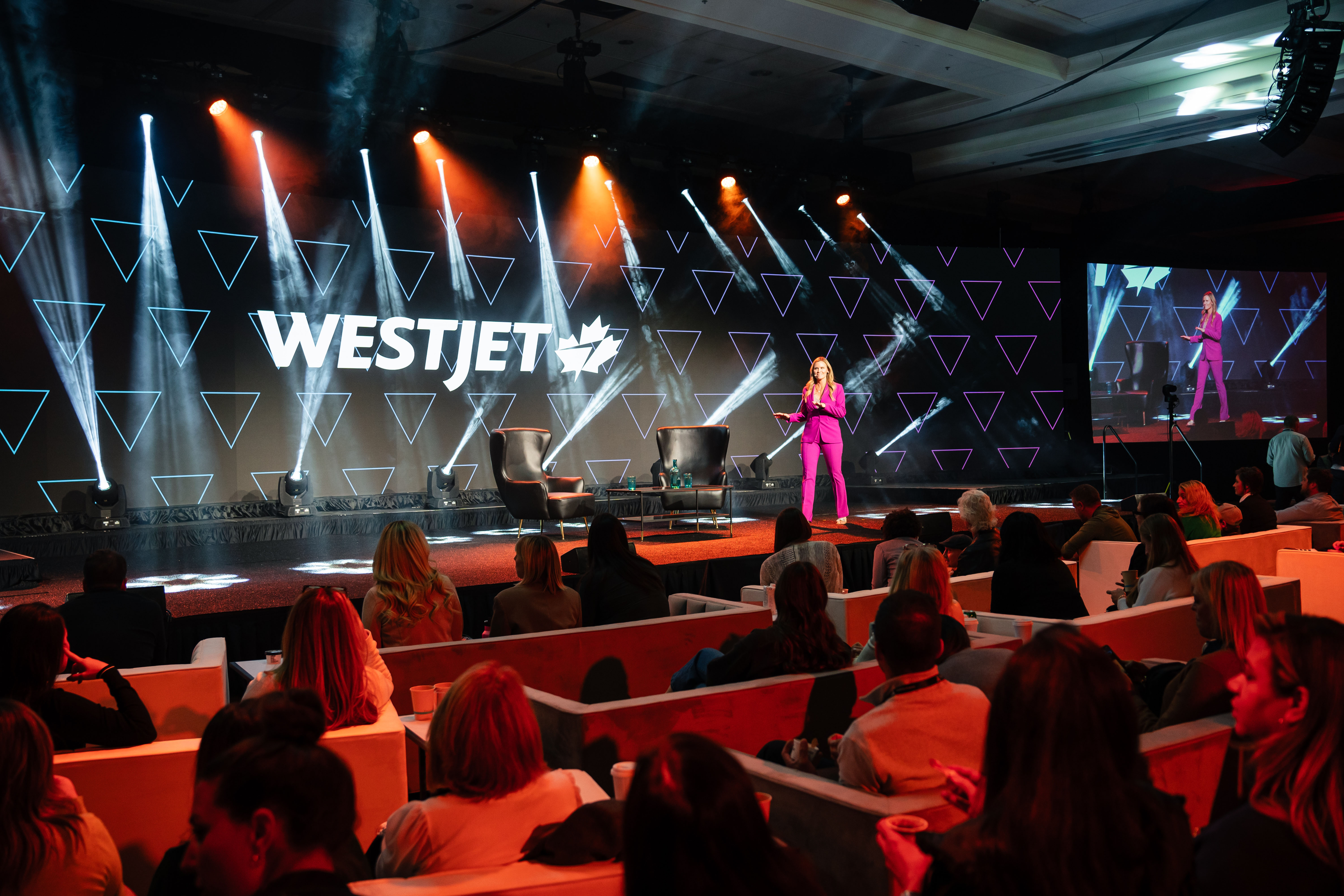 WestJet branding on an open stage with a speaker addressing an audience seated in modern booths